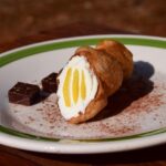 Un cannolo siciliano decorato, simbolo dell'arte culinaria del villaggio, con turisti entusiasti in background.