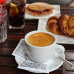 Tazza di caffè napoletano fumante su un tavolino in piazza, con sfondo di un bar storico.